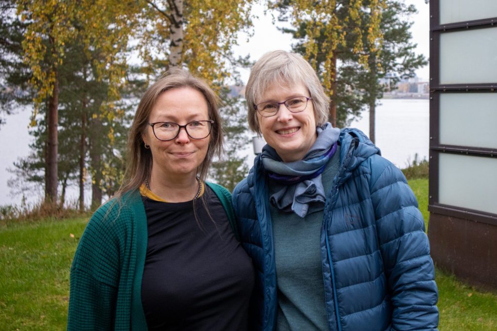 Marit Stranden og Helene Waage smiler til kamera. I bakgrunnen ser vi trær og sjø. Bildet er tatt i forbindelse med nettverksmøte for Nordic Arts & Health Research Network .