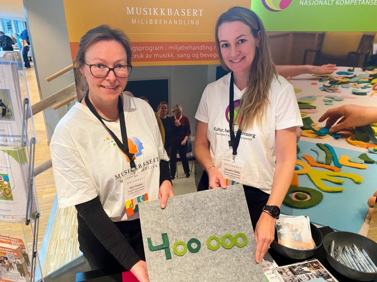 Marit Stranden og Alexandra Dahlen står på stand for Nasjonalt kompetansesenter for kultur, helse og omsorg. De smiler til kamera. Foto.