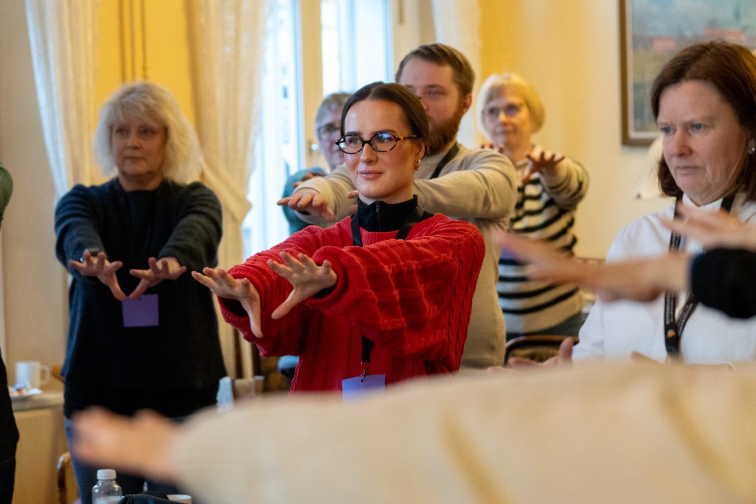 Flere personer står i et rom og strekker hendene foran seg. Foto.