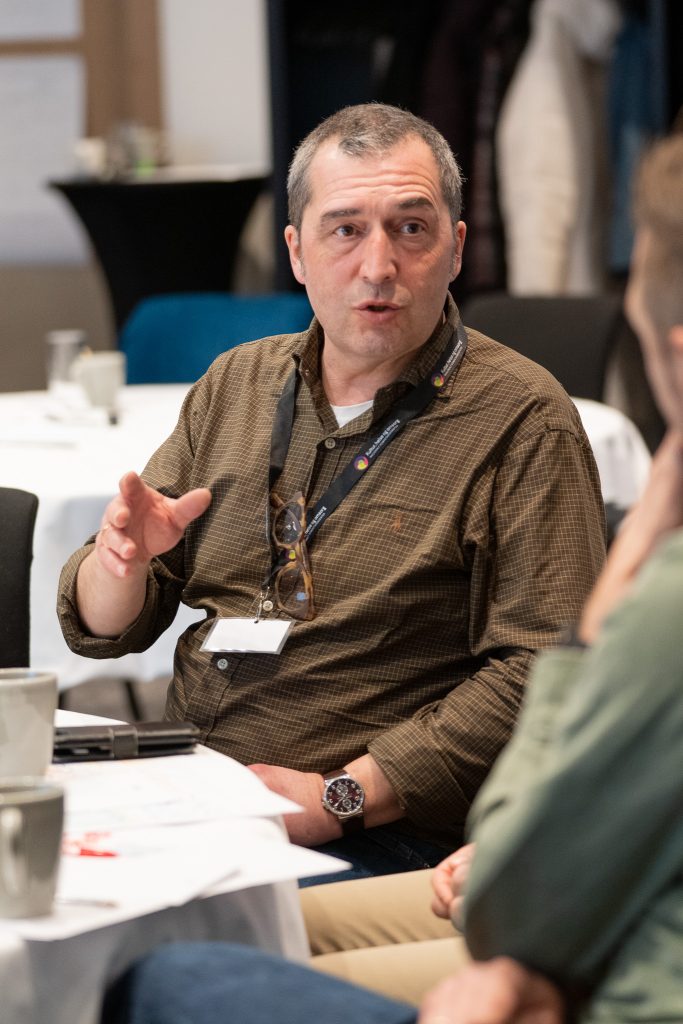 En mann sitter ved et bord sammen med andre og snakker.