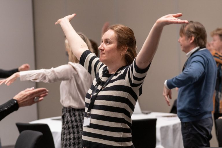 AoP_42_Foto_AlexandraDahlen En kvinne står i et rom med flere personer og strekker begge hender opp mot taket.