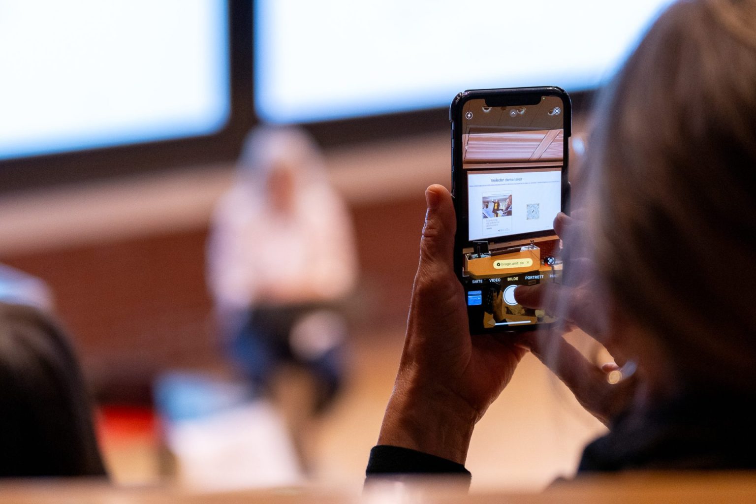En kvinne holder mobilen opp for å ta et bilde under et foredrag.