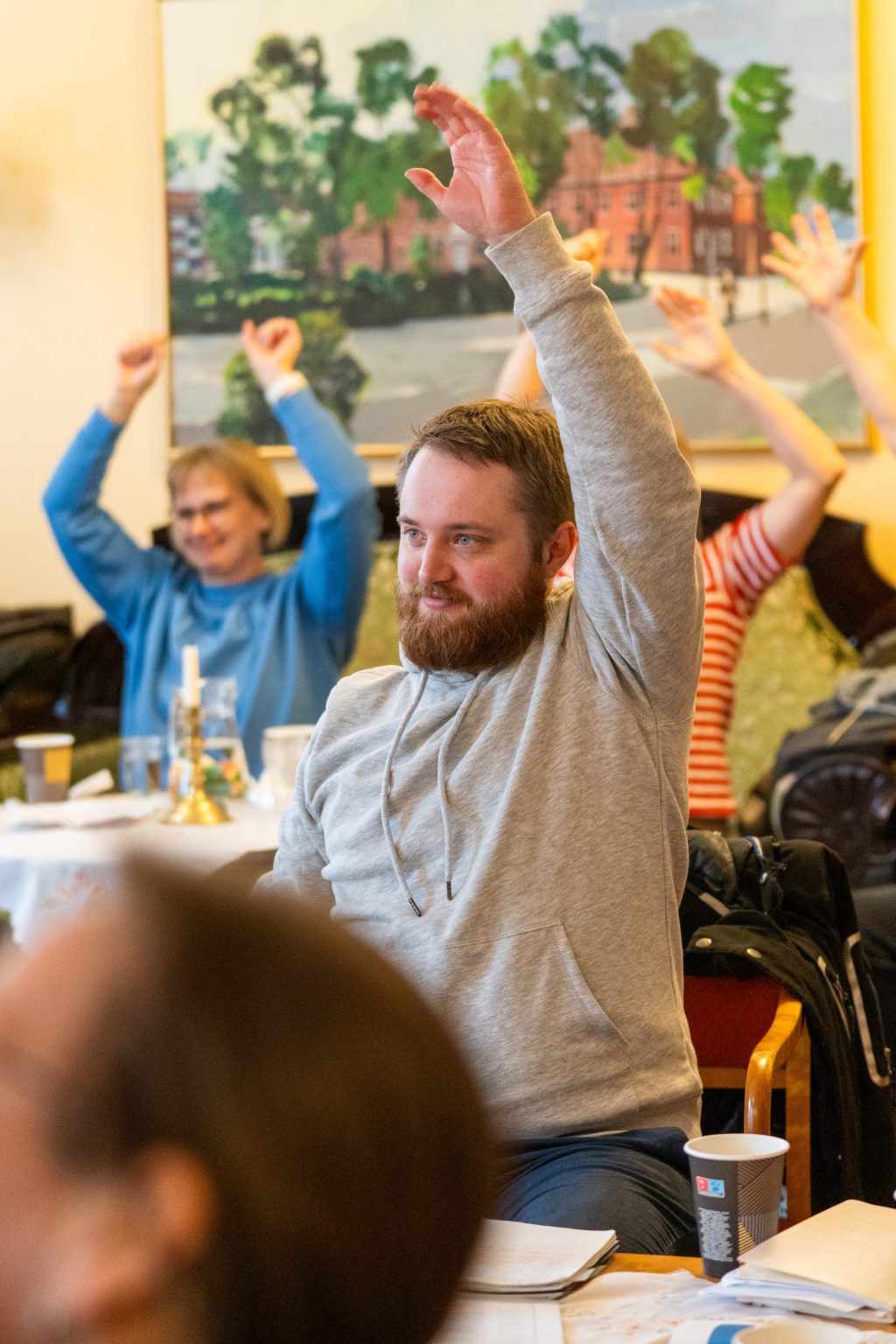 En mann sitter i et rom sammen med andre og rekker hånden i været.